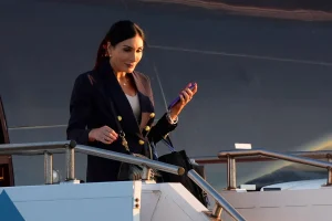 Лора Лумер на трапі літака під час приїзду в Україну, блогерка MAGA тримає телефон Альтернативний варіант (якщо потрібно точніше): Лора Лумер у темному блейзері з золотими ґудзиками стоїть на сходах літака в Україні.