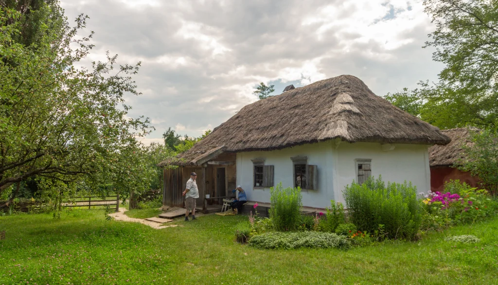 Подружжя біля білої хати з очеретяним дахом у зеленому саду